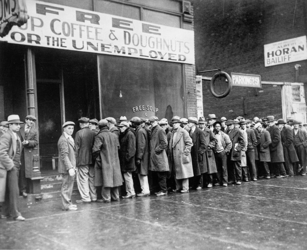 1930년대 경제 대공황의 혹독한 현실을 보여주는 사진입니다. 'FREE COFFEE & DOUGHNUTS FOR THE UNEMPLOYED' 및 'FREE SOUP'이라고 적힌 간판 아래, 수많은 실업자들이 무료 식사를 얻기 위해 길게 줄을 서 있습니다. 이는 당시의 사회적 어려움과 사람들의 고단한 삶을 상징적으로 보여줍니다. 1929 대공황 시기, 실업자들을 위한 무료 커피와 도넛, 수프를 받기 위해 길게 줄을 서 있는 사람들